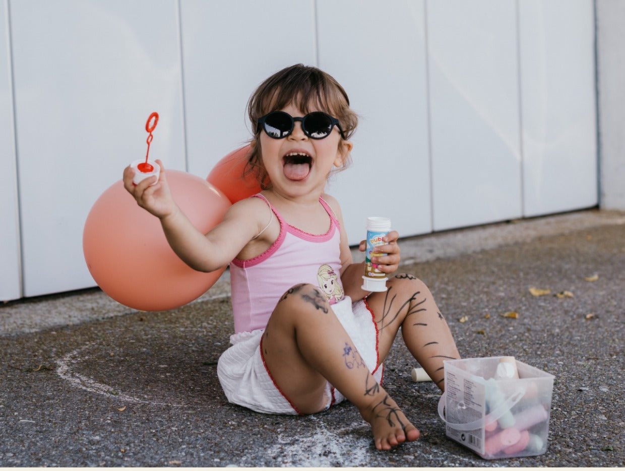 A toddler in sunglasses sitting on the pavement, covered in chalk scribbles, yelling with delight